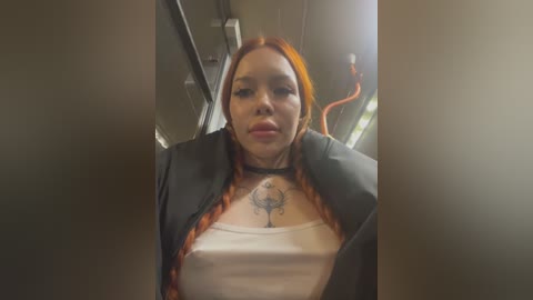 Media: Video of a woman with long, red hair and fair skin, wearing a black blazer over a white tank top revealing a tattoo of a tree on her chest, inside a dimly lit room with blurred background.