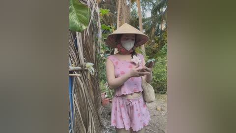 Media: Video of an Asian woman in a pink floral dress and conical hat, wearing a face mask, holding a small animal, standing among bamboo and greenery in a rural setting.