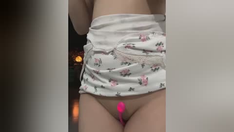 Media: Video of a light-skinned woman with a flat stomach, wearing a white floral skirt pulled up to reveal a pink vibrator inserted into her vagina. Background is a dimly lit room with a fireplace.