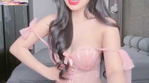 Media: Video of an Asian woman with long, wavy black hair, wearing a light pink, off-shoulder corset with frilly sleeves and a bow. She's smiling with red lipstick, in a bedroom with a gray bed and wooden wall.