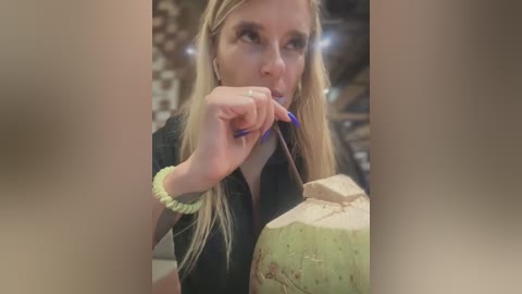 Media: Video of a blonde woman with long hair, wearing a black top and yellow bracelet, eating a green coconut with a blue nail polish-stained straw, in a dimly lit, patterned room.