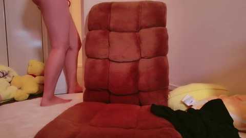 Media: Video of a plush, red, tufted chair in a cluttered bedroom. A pale-skinned person stands beside it, holding a black garment, with stuffed animals scattered around.