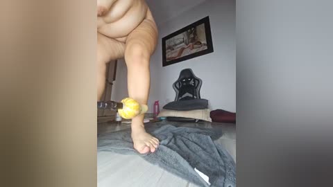 Media: A video showing a naked person with a large belly and one arm wrapped around a yellow ball, standing in a cluttered room with a gaming chair, framed painting, and scattered items.