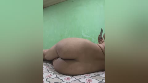 Media: A video of a naked, plus-sized woman lying on a bed with a floral-patterned sheet. Her large, round buttocks dominate the image, and she holds a phone in her right hand. The room has green-painted walls and a wooden ceiling.