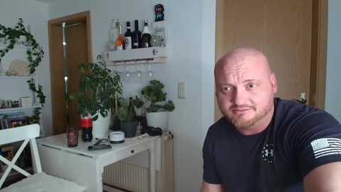 Media: A video of a bald, muscular man in a black t-shirt with a white emblem, looking perplexed, in a cluttered, plant-filled room with white shelves, wooden door, and a white chair.