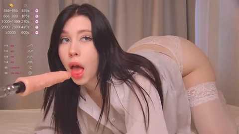 Media: A video of a pale-skinned woman with long black hair, wearing white lingerie, kneeling on a bed, licking a large, realistic dildo. The background features beige curtains.