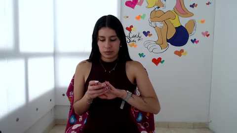 Media: Video of a Latina woman with long black hair, wearing a sleeveless maroon top, sitting on a white tiled floor, holding a small object, surrounded by colorful heart art on a white wall.