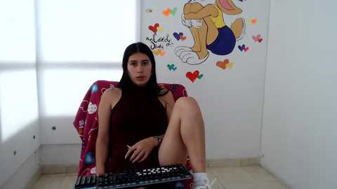 Media: Video of a young woman with long black hair in a sleeveless maroon dress, seated on a chair in a brightly decorated room. She is using a keyboard, with a Looney Tunes cartoon mural in the background.