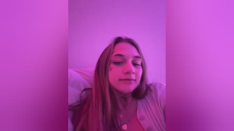 Media: Video of a young Caucasian woman with long, straight brown hair, wearing a light sweater, and lying on her side against a white wall. The image is bathed in purple light, creating a moody atmosphere.