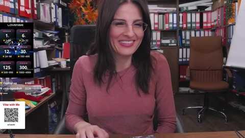 Media: Video of a smiling, middle-aged woman with long dark hair in a beige top, sitting at a desk in a cluttered office filled with colorful bookshelves and paperwork.