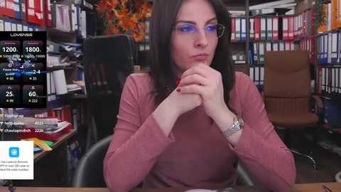 Media: Video of a woman with long dark hair, wearing glasses and a pink sweater, contemplating with her hands clasped. Background features a library with bookshelves and a desk.