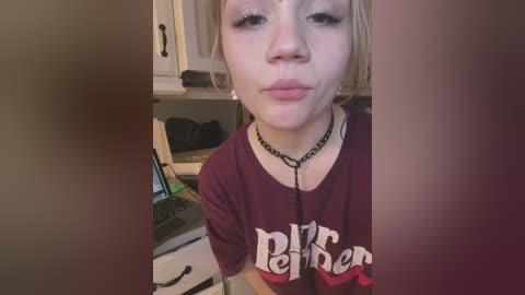 Media: Video of a young girl with light skin, blonde hair, and a maroon t-shirt with \"Rebel\" in white letters. She wears a black choker and sits in front of a desk with a laptop.