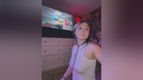 Media: Video of a young woman with fair skin and blonde hair, wearing a white tank top and black necklace, sitting in a room with a TV playing a video game.