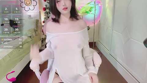 Media: Video of a young, pale-skinned woman with shoulder-length dark hair, wearing a sheer, white off-shoulder top, revealing her breasts. She is seated in a room with colorful, playful decor and a digital clock display in the background.