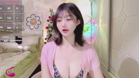 Media: A video of a young Asian woman with shoulder-length dark hair, wearing a low-cut pink top, sitting in a cozy, colorful bedroom.