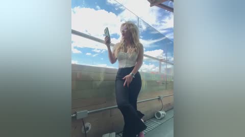 Media: Video of a blonde woman in a white lace top and black pants, holding a phone, standing on a balcony with a glass railing and a partly cloudy sky in the background.