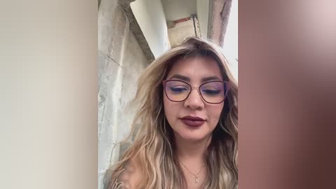 Media: A video of a young woman with long, wavy, blonde hair, wearing glasses, maroon lipstick, and a beige top, standing in front of a beige wall.