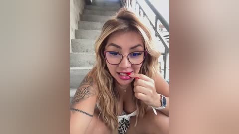 Media: Video of a young Asian woman with long, wavy blonde hair and glasses, wearing a leopard print top, licking her finger seductively while sitting on a staircase.