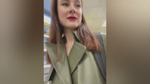 Media: Video of a young Caucasian woman with fair skin and straight, shoulder-length auburn hair, wearing a green military-style jacket, standing indoors with blurred bookshelves in the background.