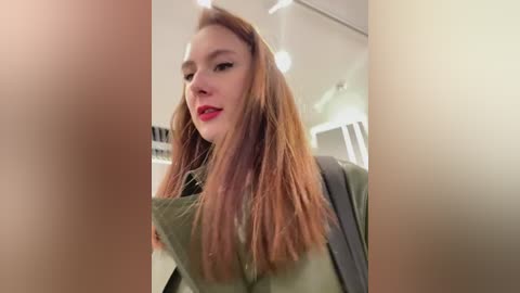 Media: Video of a young Caucasian woman with long, straight, light brown hair, fair skin, and red lipstick. She wears a green shirt, standing indoors with blurred background and a visible backpack strap.