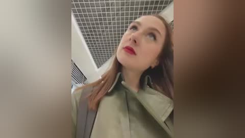 Media: A video of a Caucasian woman with fair skin, straight brown hair, and red lipstick, wearing a green coat, looking upward in a tiled, dimly lit room.