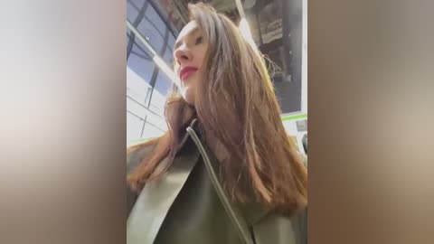 Media: Video of a young woman with long, straight brown hair, wearing a green leather jacket, captured from a low angle. The background features a modern, industrial-style interior with metallic elements and large windows.