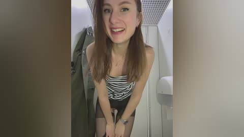 Media: Video of a young woman with long brown hair, wearing a zebra-print top and black shorts, leaning forward, smiling, in a small, brightly lit restroom with tiled walls and a white sink.