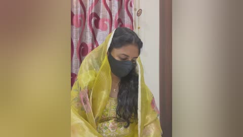 Media: Video of a woman wearing a yellow sari with floral patterns, black mask, and long dark hair, standing in a room with pink and white patterned curtains in the background.