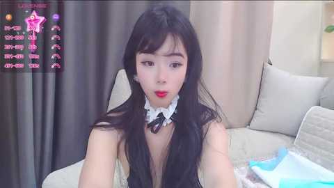 Media: A video of an Asian woman with long black hair and fair skin, dressed in a white lace choker, sitting on a beige couch. Background features curtains and a lamp.