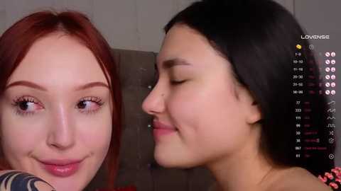 Media: Video of two young women with fair skin, one with red hair and the other with black hair, smiling and gazing at each other. Background shows a video feed with social media icons.