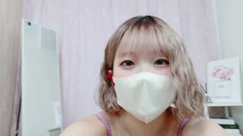 Media: Video of a young Asian woman with shoulder-length, wavy, light brown hair and fair skin, wearing a white surgical mask and a pink spaghetti-strap tank top. She has a neutral expression and is indoors, with a white curtain and a white cabinet in the background.