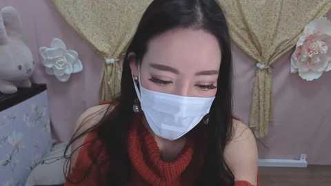 Media: Video of a young Asian woman with long black hair, wearing a white face mask, red sweater, and dangling earrings, seated in a room with pink walls, floral curtains, and a teddy bear.