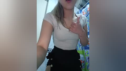 Media: Video of a slender, light-skinned woman with shoulder-length blonde hair in a white t-shirt and black high-waisted skirt, standing in a brightly lit bathroom with a colorful shower curtain.