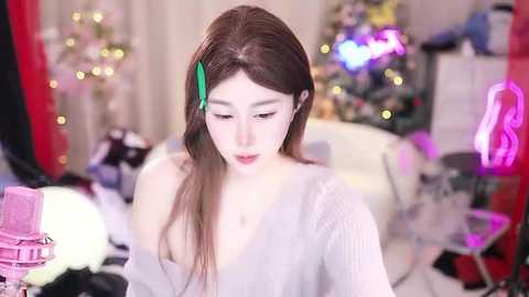 Media: Video of an Asian woman with fair skin, long brown hair, and a green hair clip, wearing a gray, off-shoulder sweater, standing in a softly lit, festive room with a Christmas tree and pink gaming chair in the background.