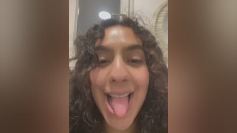 Media: Video of a young woman with curly, dark hair, medium brown skin, and a wide smile, sticking out her tongue. The background features a bathroom with a shower curtain and a white wall.