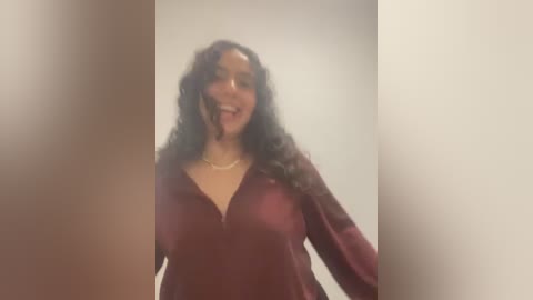 Media: Video of a smiling woman with medium skin tone, curly black hair, wearing a deep burgundy V-neck top, against a plain beige wall.