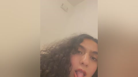 Media: A video of a young woman with curly hair, light brown skin, and expressive eyes, playfully sticking out her tongue. The background is a plain, beige wall with a small vent in the top left corner.