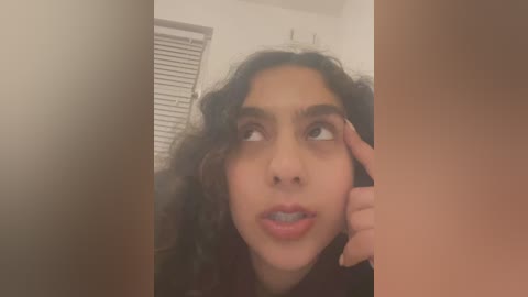 Media: A blurry, close-up video of a young woman with curly dark hair, light skin, and a confused expression, touching her forehead. The background shows a white wall and closed blinds.