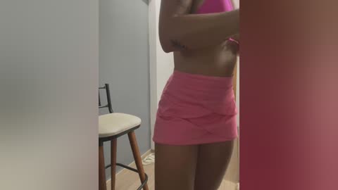 Media: A video of a slender woman in a tight pink mini-skirt and matching crop top, standing in a narrow hallway with a white chair and a light blue wall.