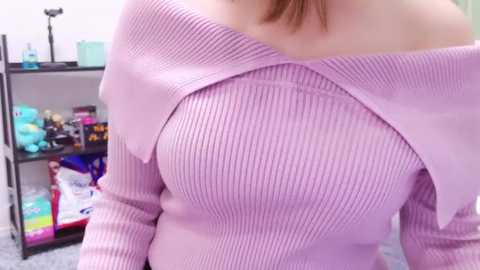 Media: Video of a woman wearing a pale pink, ribbed sweater with a dramatic, asymmetrical neckline. Background features a cluttered shelf with various items, including snacks and a camera.