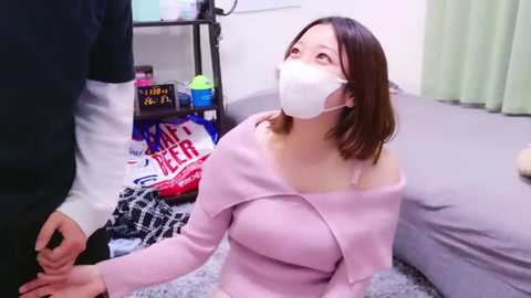 Media: Video of a young Asian woman with shoulder-length brown hair, wearing a white mask and a light pink off-shoulder sweater, kneeling on a gray carpet in a cluttered living room.