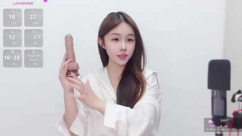 Media: Video of an East Asian woman with straight, dark hair, wearing a white robe, holding a realistic flesh-colored dildo, surrounded by temperature and size charts, in a minimalist room.