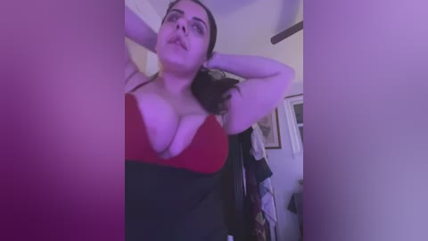 Media: A low-angle video of a plus-size woman with dark hair, wearing a red and black bra, standing in a dimly lit room, with purple lighting.