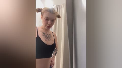 Media: A video of a young, pale-skinned woman with a slim build and light brown hair styled in two pigtails. She wears a black sports bra and has several tattoos visible on her chest and arms. The setting is a minimalist bathroom with beige walls and a white curtain.