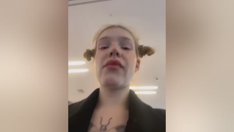 Media: Video of a young woman with fair skin and blonde hair styled in two small buns. She has a neutral expression, wearing a black jacket over a dark top with a visible tattoo. The background is a plain, light-colored room with fluorescent lighting.