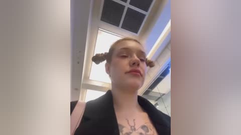 Media: Video of a young woman with fair skin and blonde braids, wearing a black blazer, standing indoors. She has a tattoo on her chest. Background features a grid-patterned ceiling and fluorescent lights.