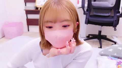 Media: Video of an Asian woman with light brown hair and light skin wearing a white face mask and a white sweater. She is sitting on a white floor, with a pink basket and office chair in the background.