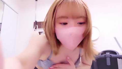 Media: A video of a young Asian woman with light skin and straight, shoulder-length blonde hair, wearing a light blue bra, covering her mouth with a pink mask. She is leaning forward, hands on the floor, in a bathroom with white walls and a black chair.