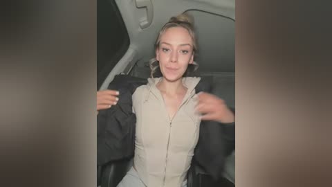 Media: A video of a young woman with light skin, blonde hair tied in a bun, wearing a beige zip-up jacket and gray pants, sitting in a car.