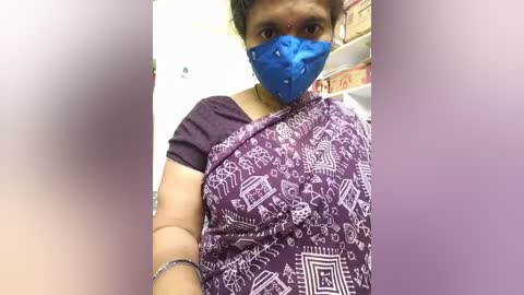 Media: Video of an Indian woman wearing a blue face mask, purple sari with white geometric patterns, and a dark purple short-sleeved blouse. She stands indoors with a blurred background featuring shelves.